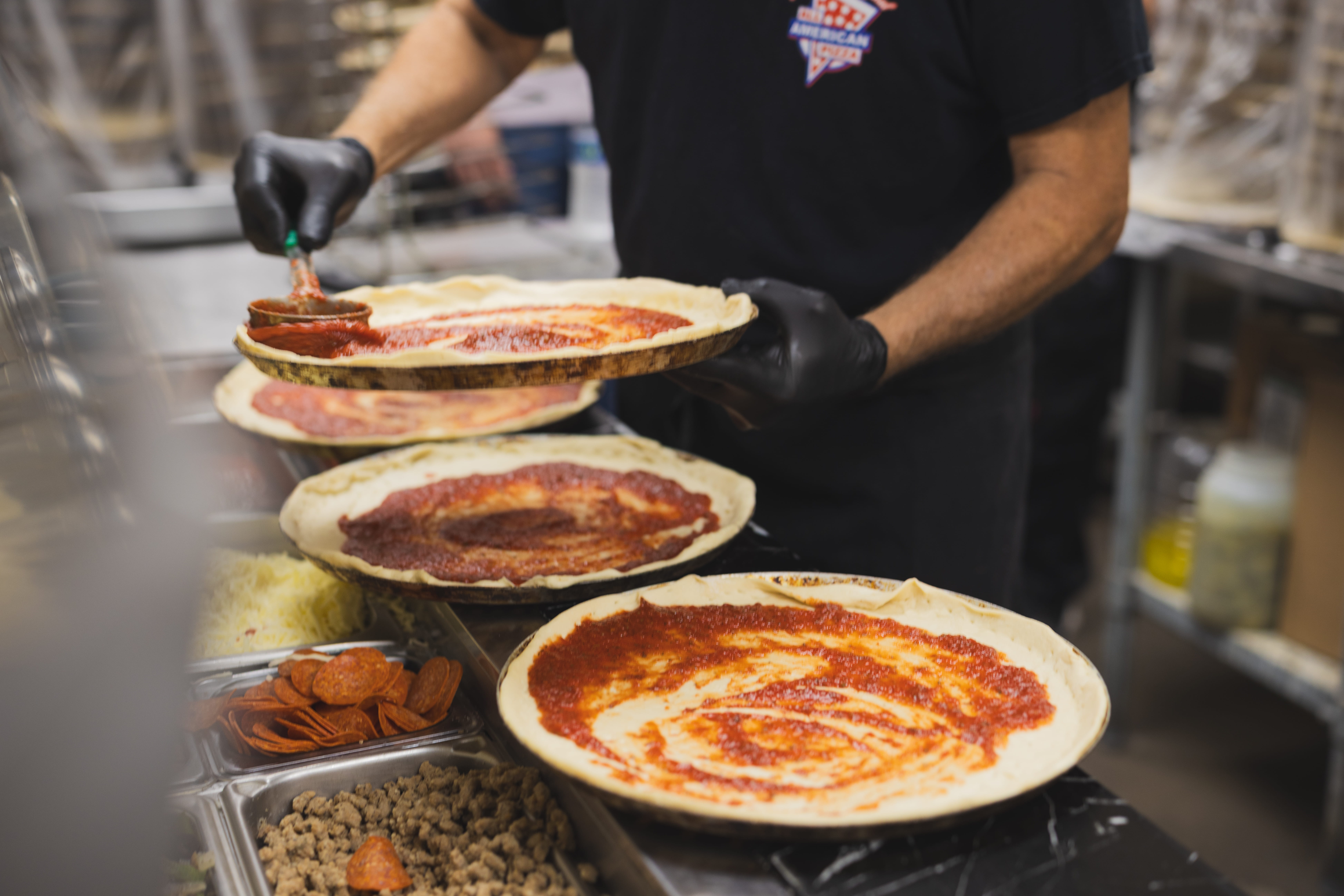 Pizza preparation in a busy kitchen: a cook wearing black gloves spreading tomato sauce on round pizza crusts lined up on the prep station, with pepperoni toppings visible on some pies.
