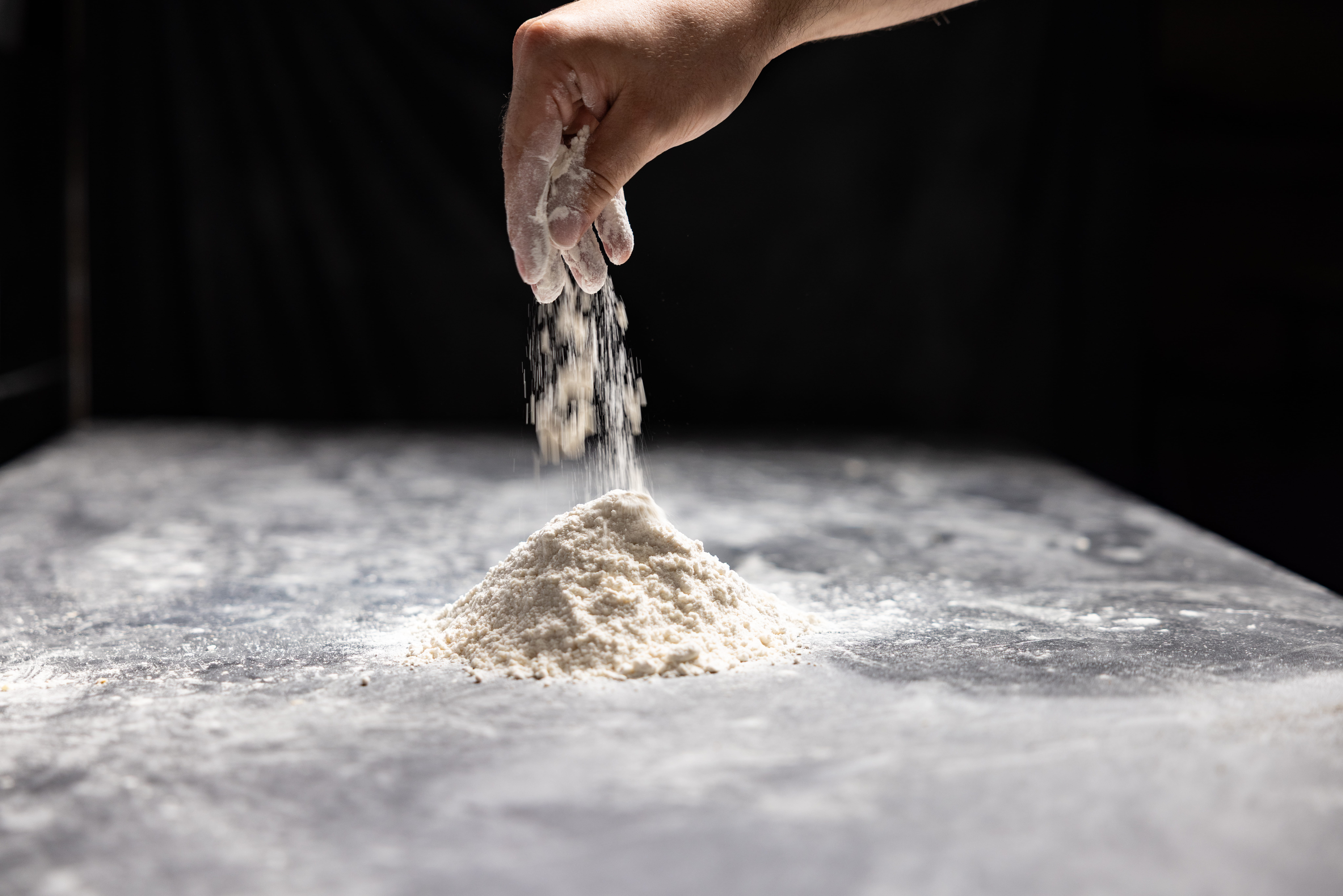 Close-up of a hand dusting flour onto a mound of dough on a floured surface with a dark background