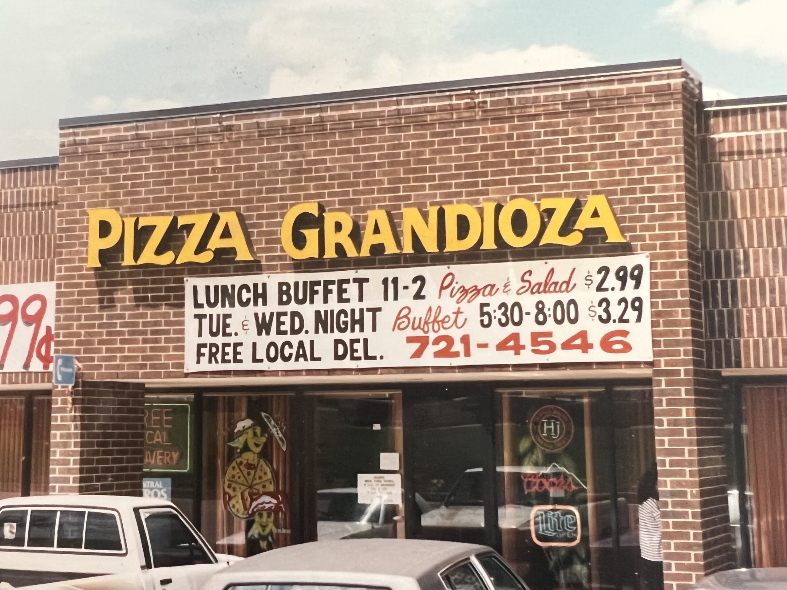 Brick storefront with a large yellow 'Pizza Grandioso' sign on a brick wall and white menu banners beneath